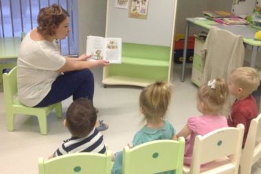 Воспитательница показывает детям, сидящим на стульях, картинку в книжке