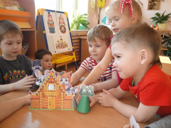 Три мальчика и девочка разыгрывают сказку «Теремок» на столе с картонными персонажами