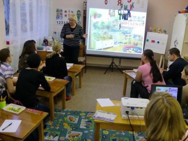 Воспитательница презентует свой опыт на экране перед сидящими за столами коллегами