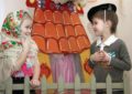 Мальчик и девочка в платке играют сценку на фоне декораций: домика и забора