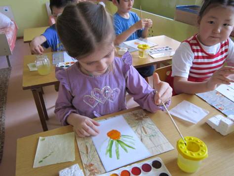 Дети сидят по двое и рисуют красками, девочка дорисовывает зелёные штрихи к оранжевому овалу в центре листа