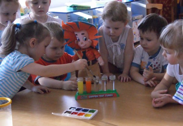 Дети стоят вокруг стола с водой и красками, рядом с ними кукла Незнайка