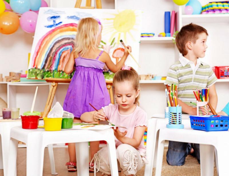 Девочка, стоящая спиной, рисует солнце на мольберте, девочка справа рисует красками, мальчик слева смотрит в сторону