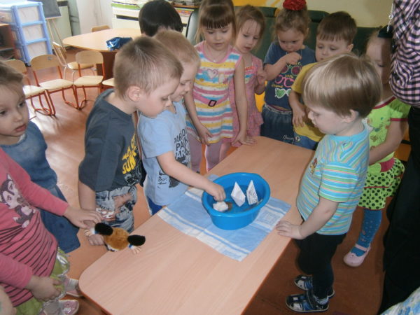 Группа детей наблюдает за плавающими в синем тазу бумажными корабликами