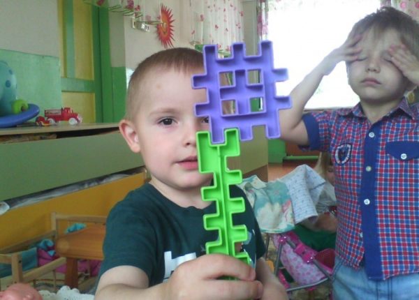 Мальчик держит сложенные детали конструктора, на заднем фоне другой мальчик закрыл глаза руками