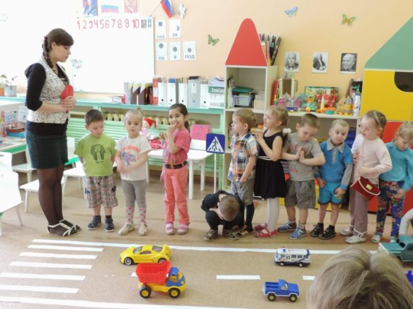 Воспитательница что-то говорит детям, стоящим цепочкой перед имитацией пешеходного перехода
