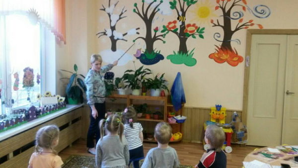 Воспитательница показывает на стену с макетами деревьев в разное время года, дети стоя слушают