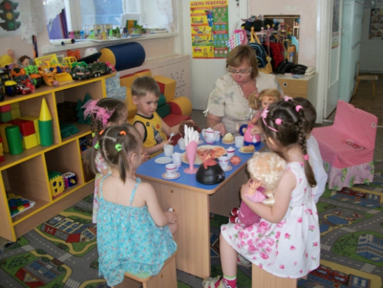 Воспитательница сидит в окружении детей за накрытым игрушечной посудой столиком