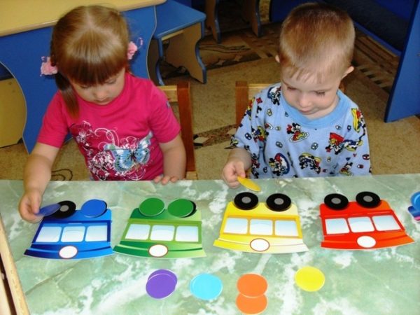 Девочка и мальчик закрывают цветными кругами колёса разноцветного картонного поезда