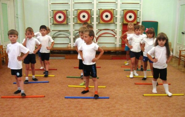 Дети в спортивном зале катают гимнастические палки ногами