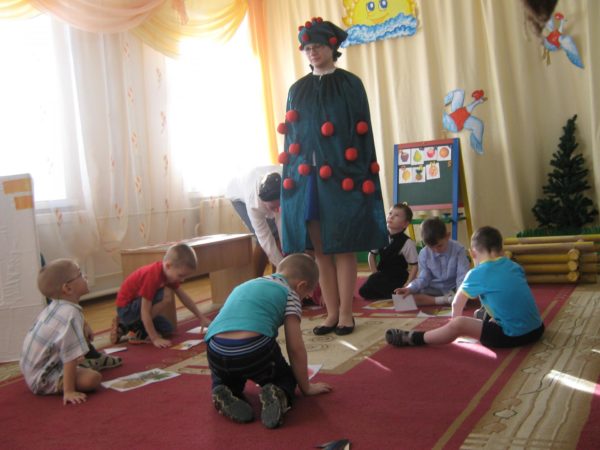 Дети сидят в кругу на ковре, перед ними лежат картинки с фруктами, в центре стоит педагог в костюме яблони