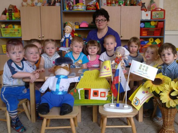 Педагог и малыши сидят за столом, перед ними — куклы и флаги