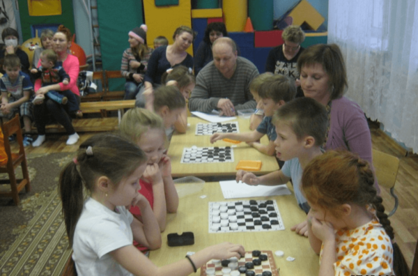 Дети играют в шашки в ходе турнира, родители наблюдают